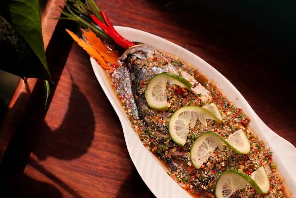 A whole fish dressed with chopped chili and herbs and slices of lime, on a long white plate.