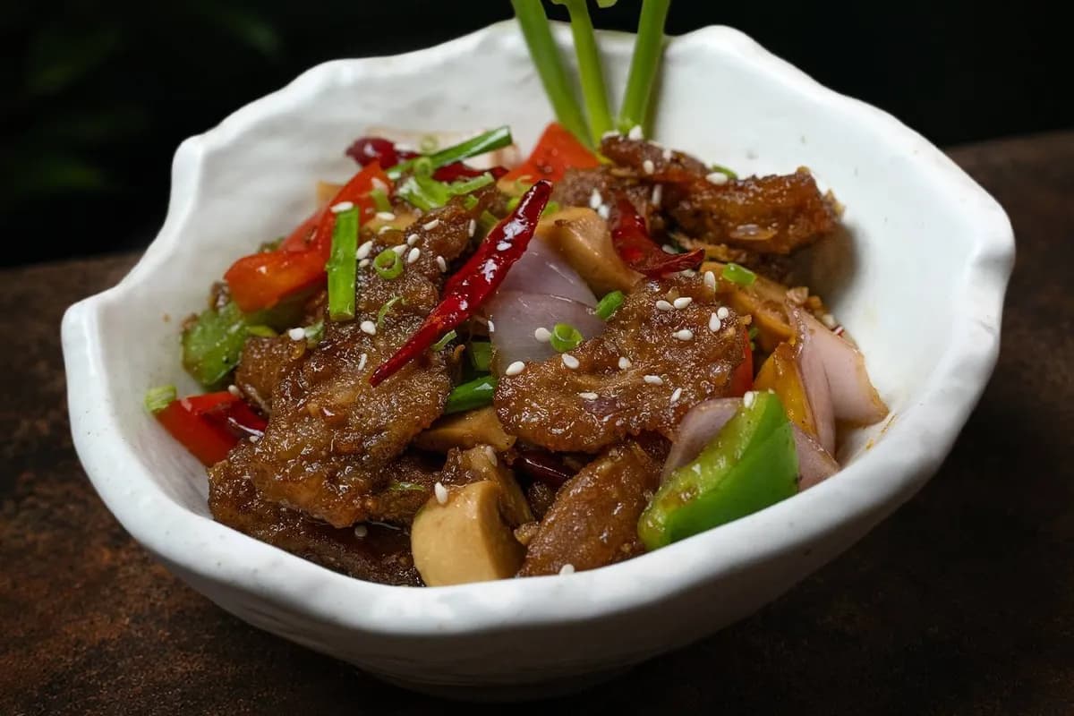 Slices of meat stir-fried with peppers, onion and dried chili, sprinkled with sesame, in a white bowl.
