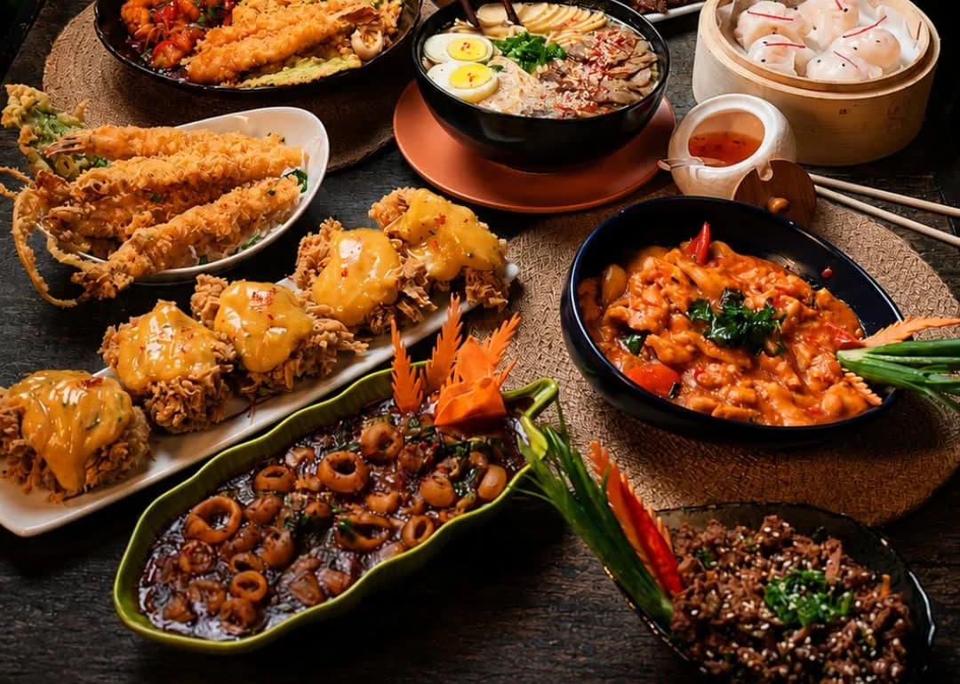 A dark table crowded with dishes: fried prawns, a bowl of ramen, steamers of dumplings, squid rings in sauce and a red curry.