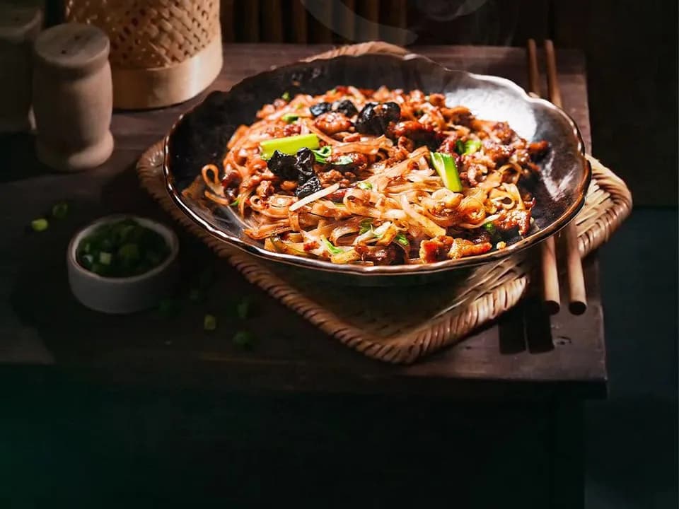 Pad thai noodles with greens and mushrooms in a dark bowl on a woven tray.