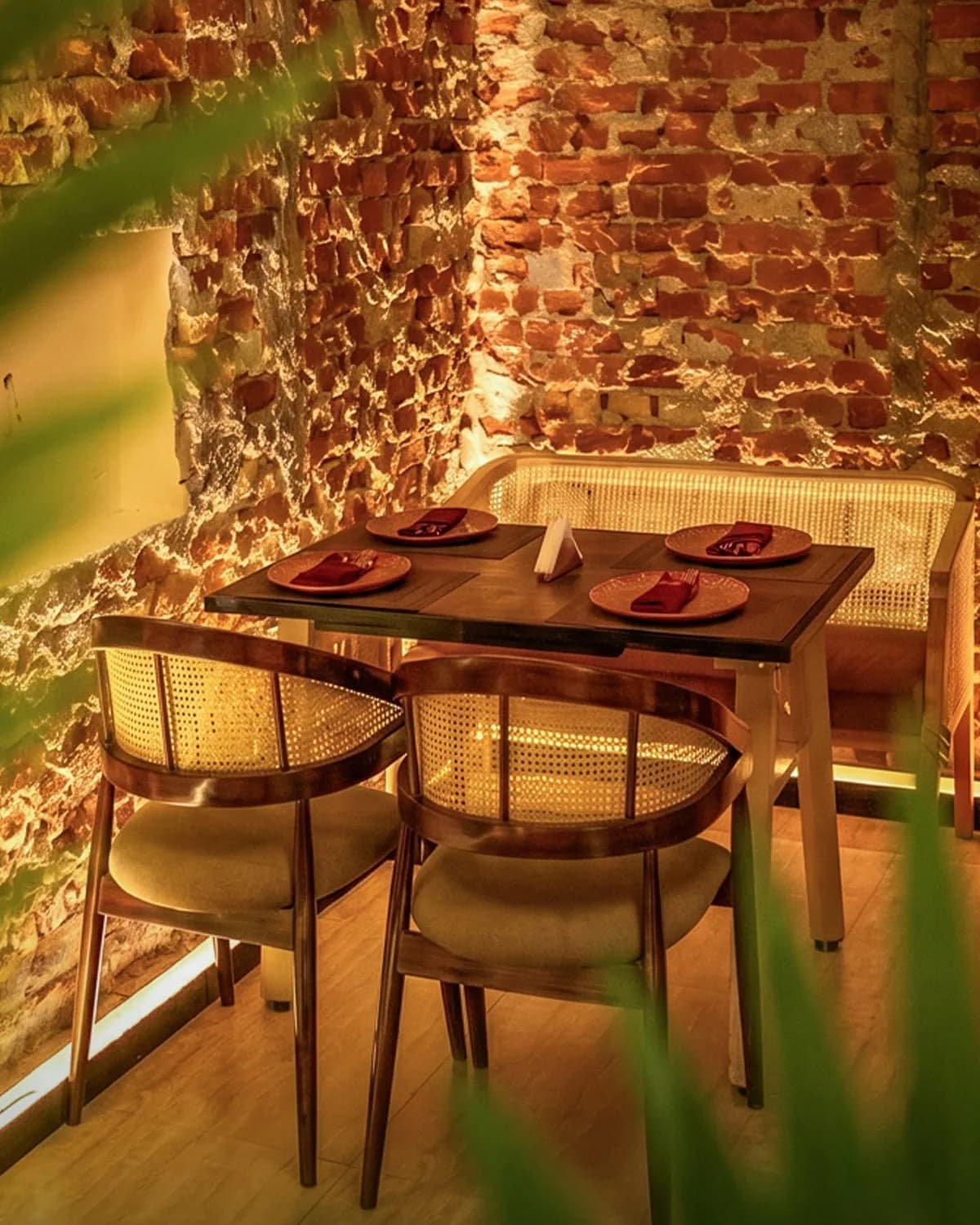 A table set for four with cane-backed chairs and a bench, in a corner of warmly lit brick walls.