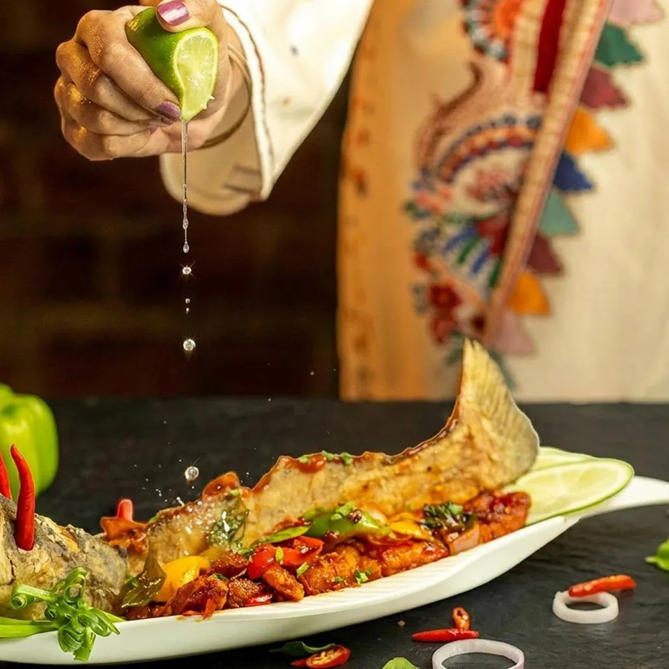 A hand squeezes lime over a whole fried fish dressed with chili and herbs on a long white plate.
