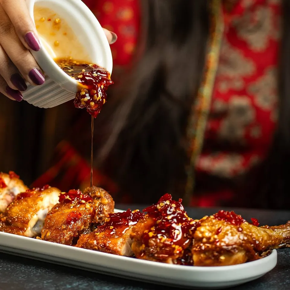 Chili sauce poured from a small white cup over glazed chicken on a long plate.