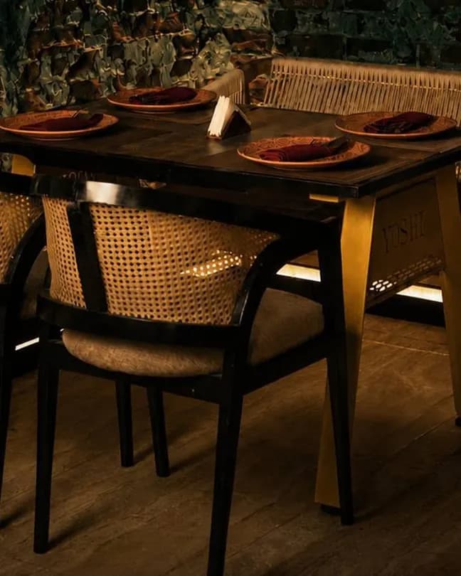 A dark wooden table laid with plates and red napkins, with cane-backed chairs and a woven bench by a brick wall.