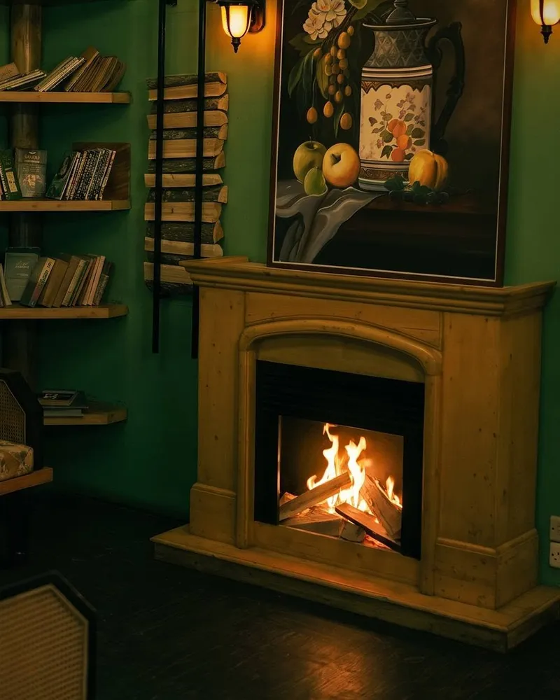 A green-walled corner with a lit fireplace under a still-life painting, wall lamps and shelves of books.