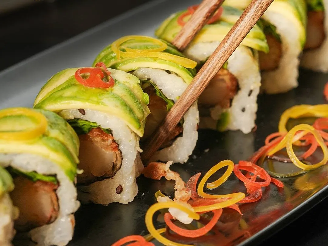 Rolls topped with sliced avocado and curls of vegetable, with chopsticks lifting one piece.