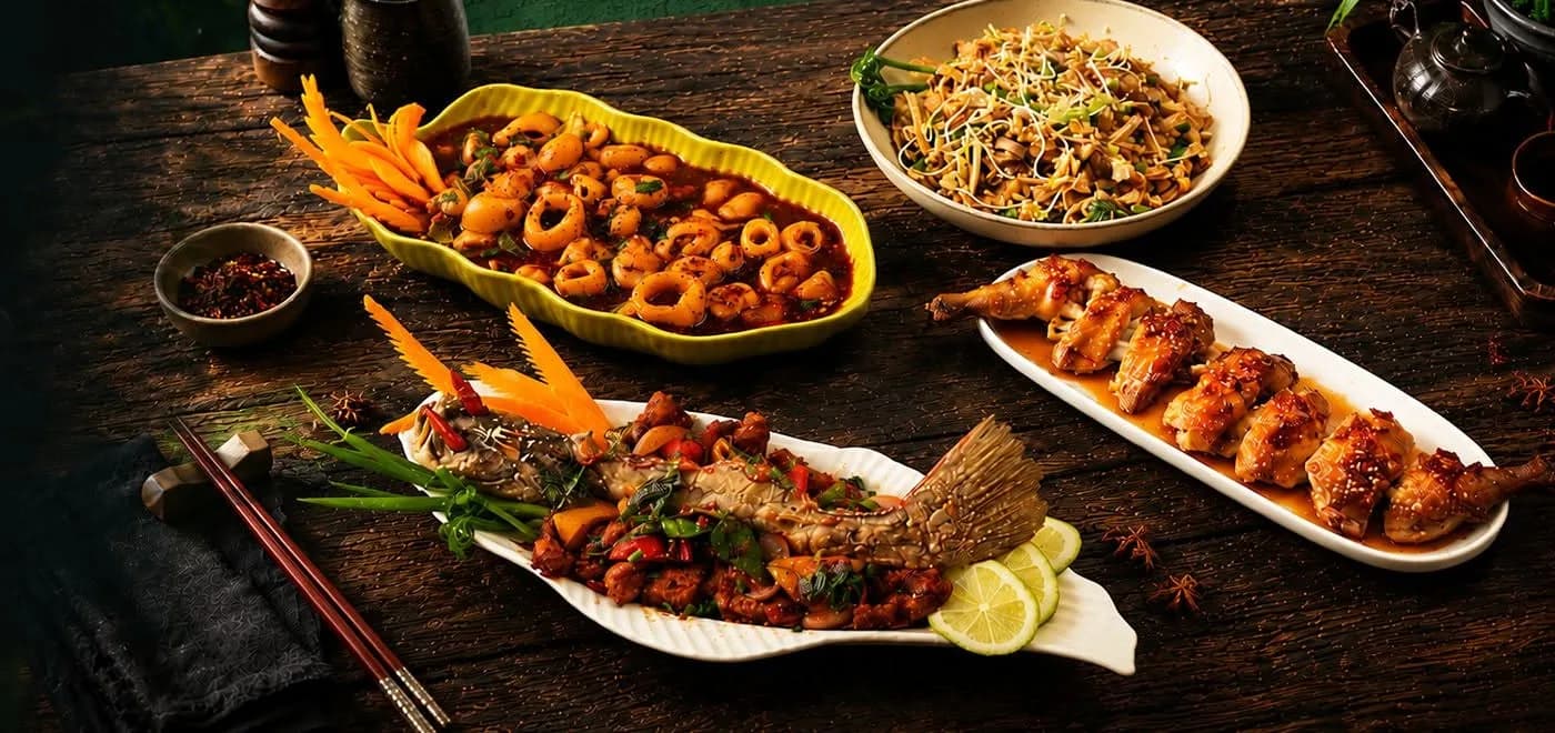 A wooden table laid with four dishes: squid rings in a dark sauce, a whole fish with lime, glazed chicken and a bowl of noodles.